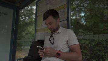 Young caucasian man at the bus stop waiting for the bus in the hands of a mobile phone sends messages. Theme of urban transport and mobility, traveling around Europe in comfortable transport. video