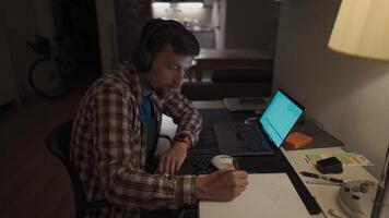 Student does homework onlaptop at home and listens to music on headphones. Man studies online at homeroom and listsens webinar and takes notes in notebook with pen. Audio course, e-learning education. video