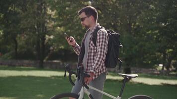 A male cyclist walks his bicycle through a park and uses a smartphone in sunny weather in Germany. A man is pushing a bicycle and surfing the Internet on his phone in a city park in Europe video