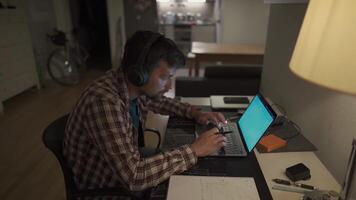 A male student studying on his laptop with headphones and taking notes in his notebook with a pen at home in the evening. The male works at the computer late at night with headsets in the apartment. video