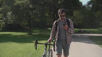 A male cyclist walks his bicycle through a park and uses a smartphone in sunny weather in Germany. A man is pushing a bicycle and surfing the Internet on his phone in a city park in Europe video