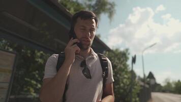 Man late wait for transport at bus station, nervous, and calls to report being late in summer in Germany. Delay. Schedule of route. Male speaking on mobile phone while awaiting delayed vehicle. video