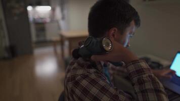 Tired male student studying on laptop at home until late and has neck pain and massages himself with percussion gun. Neck and back pain. Overworked man uses pistol massager to relieve muscle tension video