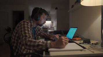 Student does homework onlaptop at home and listens to music on headphones. Man studies online at homeroom and listsens webinar and takes notes in notebook with pen. Audio course, e-learning education. video