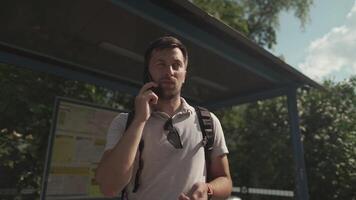 Man checks time on wristwatch while waiting for bus at stop, calls to warn of delayed appointment, male communicates on cell phone while delaying tranport in Germany. An irritated male is late. video