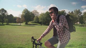 A male with a backpack on a bicycle ride in the park on his cycle talking on the phone in sunny weather. A bicyclist rides his bike in the park and talks on the telephone with a rucksack. video