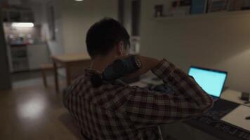 Tired male student studying on laptop at home until late and has neck pain and massages himself with percussion gun. Neck and back pain. Overworked man uses pistol massager to relieve muscle tension video