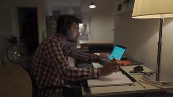 Student does homework onlaptop at home and listens to music on headphones. Man studies online at homeroom and listsens webinar and takes notes in notebook with pen. Audio course, e-learning education. video