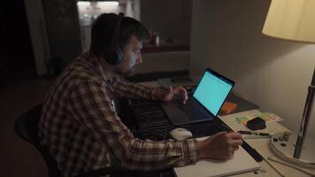 Focused man in headphones with a laptop late at night at home, taking notes, student learning a language, watching an online webinar, hearing an audiocourse, the concept of e-education. Homework. video