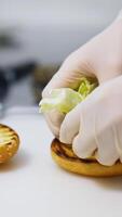 Hands in latex gloves put lettuce on top of fried bun. Making burger on white table. Fast food concept. Vertical video