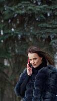 luxe fille avec une téléphone dans le parc. portrait de attrayant Dame dans fourrure manteau en marchant dans le parc et parlant le mobile téléphone. lent mouvement. verticale video