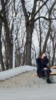 Girl sits on bench in the park. Beautiful young woman in fluffy coat sitting alone outdoors on the leafless trees background. View from below. Vertical video