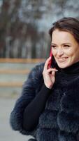 Smiling woman talking the phone. Cheerful girl in fur coat standing in the park and speaks on the mobile phone. Slow motion. Vertical video