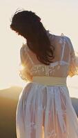 Dark-haired woman in transparent white dress standing high in the mountain. Unrecognizable lady stands her back to the camera enjoying setting sun. Vertical video