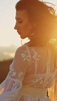 Long-haired lady wearing white dress standing high in the mountains. Lady is turning around at the backdrop of shining bright setting sun. Vertical video