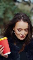 Portrait of a smiling woman. Attractive brunette girl in fur coat stands with plastic cup of coffee in the park and looking on camera. Slow motion. Vertical video