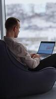 Working man sits with his back to the camera typing on his laptop. Male employee building charts on computer. City panorama in blur at backdrop. Vertical video