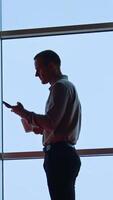 Man sitting at the desk, looking at his phone, talking and smiling to somebody in the room. Low angle view. Panoramic windows at backdrop. Vertical video