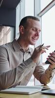 Man sitting at the desk, looking at his phone, talking and smiling to somebody in the room. Low angle view. Panoramic windows at backdrop. Vertical video