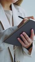 Lady in light jacket standing at the window and holding paper notebook. Woman making notes in her diary. Close up. Vertical video