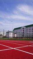 Approaching a soccer field surrounded by fence in front of the big pink building. Red running track at the foreground. Low angle view. Vertical video
