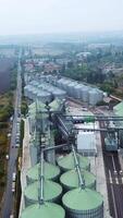 The territory of a huge plant divided by railways. Silo tanks of granary elevator at the backdrop of farmlands and countryside. Vertical video
