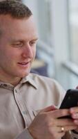 Caucasian man sits comfortably in a chair, talking and looking at his phone. Rest from work during lunch break. Blurred backdrop. Vertical video