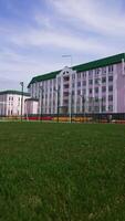 Red running track, green grass and football field located in front of the beautiful pink building. Low angle view. Blue sky at backdrop. Vertical video
