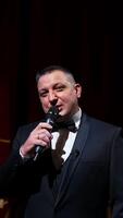 Speaker on stage. Male host in elegant costume speaking into a microphone looking on camera on the background of red curtains in the theater. Slow motion. Vertical video