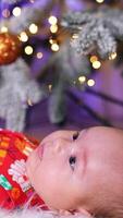 Beautiful Caucasian child lies resting under Christmas tree. Little newborn kid start to wave his tiny hands and feet. Side view. Vertical video
