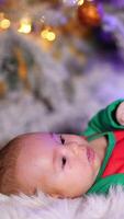 Tiny infant baby boy in green costume of elf on white fluffy plaid. Beautiful kid lying at the backdrop of Christmas tree in blur. Vertical video