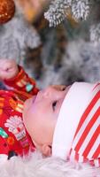 Nice little toddler in red outfit and red stripy cone hat lies on a soft white plaid. Adorable baby in costume under Christmas tree. Blurred backdrop. Vertical video
