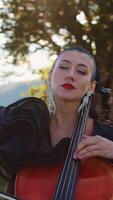 Mid-aged young woman with makeup and nice black dress performs cello music. Lady closed her eyes enjoying music. Nature backdrop in blur. Vertical video