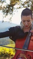 Violoncello musician with bright makeup and long earrings playing instrument. Lady stops to fix the earring and goes back to playing. Nature backdrop. Vertical video