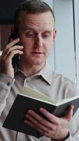 Man in grey shirt makes phone call. Male employee holds a paper notebook in front of him and speaks on the phone. Vertical video