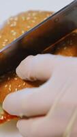 Hands in gloves of a cook making burger. Chef cutting ready hamburger into halves with a sharp knife on table. Close-up. Vertical video