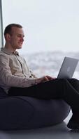 Middle-aged man sitting in chair works on his laptop. Male employee working in comfortable office atmosphere. Side view. Panoramic windows in blur at backdrop. Vertical video