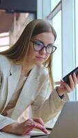 Long-haired woman in glasses uses phone and laptop at once. Lady stands above the desk and types something on computer. Vertical video