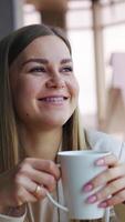 Happy smiling lady enjoys rest and tea during lunch break. Young woman looks dreamily into the window. Vertical video