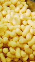 Worker's gloved hand moves a mass of corn sticks with spatula. Crispy snacks close up at conveyor belt. Vertical video