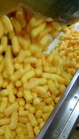 Lots of yellow crispy corn sticks fall on the conveyor line. Factory worker pushes snacks along the belt. Food factory production concept. Vertical video
