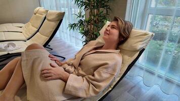 A Caucasian woman in a spa robe relaxes on a lounge chair, embodying self care and relaxation video