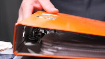 close-up Office worker stacks folders with documents. Office worker is consulting documents inside an orange binder, potentially reviewing important paperwork or preparing for a meeting video