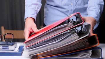Office worker is putting a red ring binder on top of a stack of colorful ring binders full of paperwork, on a white desk. office worker puts a stack of folders with documents on his desk video