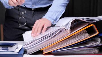 Businessman searching information holding eyeglasses and pointing at documents in folders. office worker searches for important information in folders, surrounded by paperwork and office supplies video