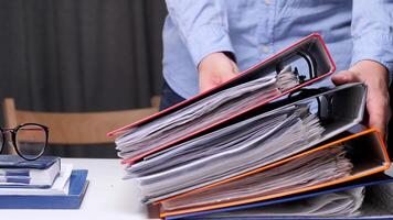 concept of storing documents in the archive. A large stack of official documents. Office worker is placing a tall stack of colorful ring binders full of paperwork on a white desk, suggesting a busy video