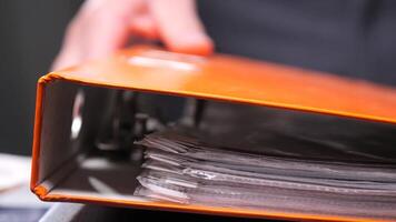 Office worker is consulting documents inside an orange binder, potentially reviewing important paperwork or preparing for a meeting video