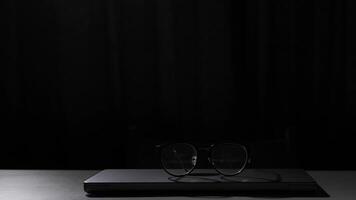 Eyeglasses resting on closed laptop on white table with chair in background. Eyeglasses are resting on a closed, silver laptop computer on a white table with a wooden chair in the background video