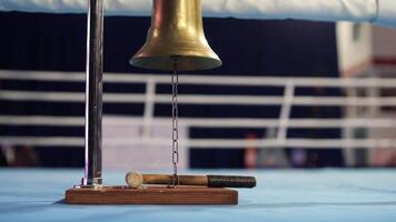 Shiny brass boxing bell hangs from a chain, with a mallet on a wooden platform by the ring ropes. The blue bell contrasts with warm wood tones, capturing the excitement of boxing. Concept of start video