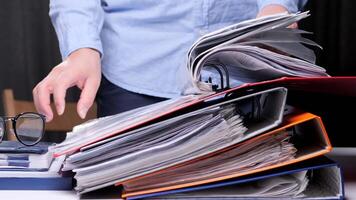 office worker studies a contract, document in the archive, stack of binders. Office worker is searching through a large stack of colorful binders and files, possibly looking for important document video